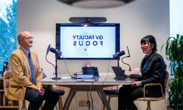 Two people smile while sitting on chairs. Microphones are in front of them. A screen with the words, "GV Faculty Focus" is in the background.