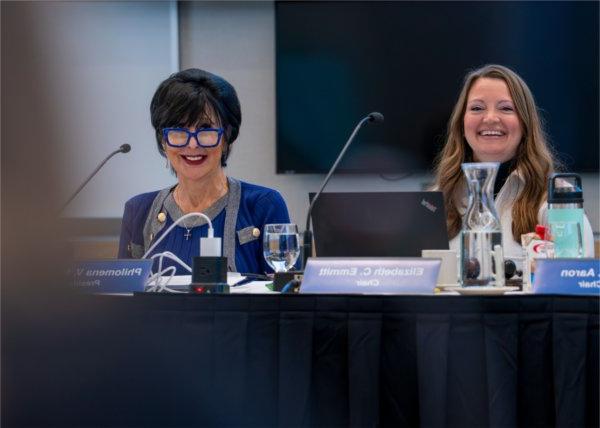 Two people seated in front of microphones smile during a meeting.